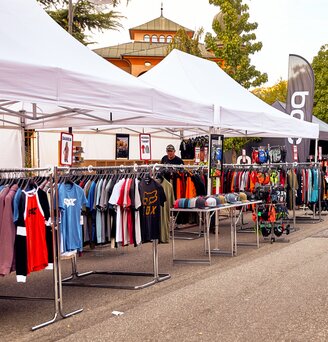 The white promotional tents serve as a sales tent at an event. Various t-shirts hang under the folding gazebo.