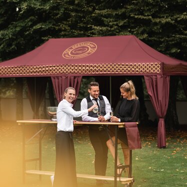 Elegant folding gazebo 8 x 4 m burgundy with gold decorations and decorative pole-covering curtains. The gazebo stands in a meadow and is used for the reception of an elegant event while people drink sparkling wine served by a waitress