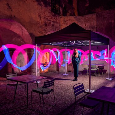 The black folding gazebo stands in a courtyard. Under it stands a young woman. Around the folding tent there are light strips in pink and blue.